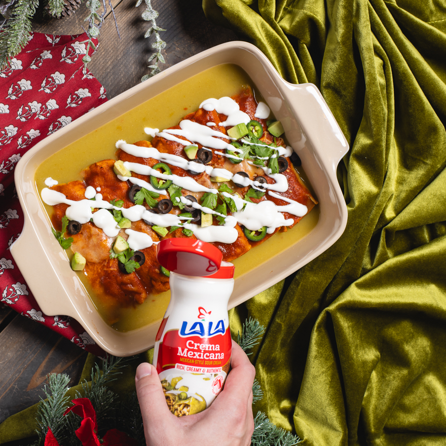 A baking pan of enchiladas being topped with Lala crema Mexicana