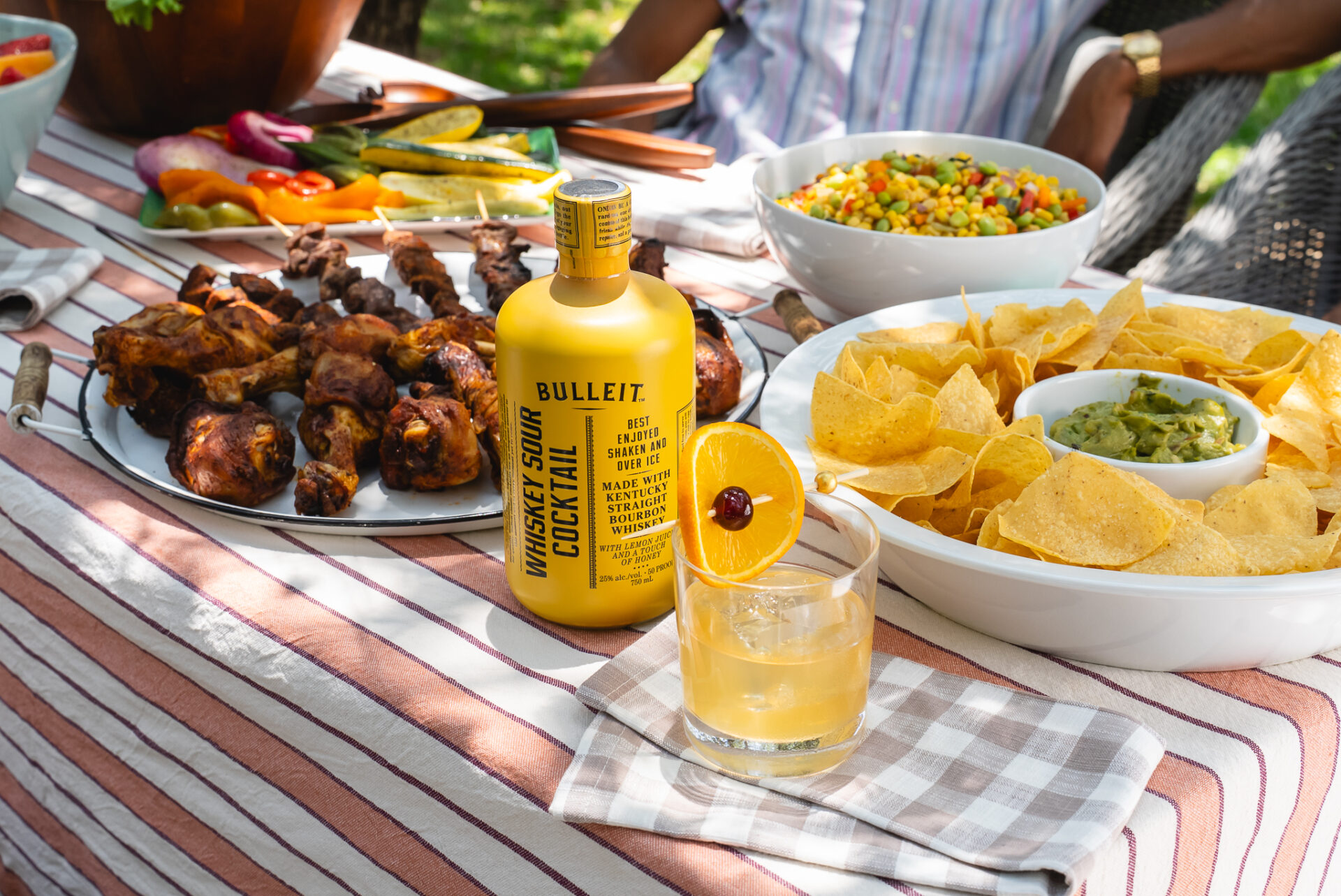 A bottle of The Cocktail Collection Bulleit whiskey sour on a summer table with grilled food